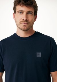 Marinblå bomulls t-shirt med rund hals, korta ärmar och en grå fyrkantig logotyp på vänster bröstområde. Slät textur.