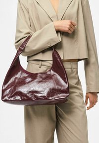 Osoba u bež odijelu drži veliku sjajnu burgundsku kožnu torbu na lijevom laktu uz bijelu pozadinu.