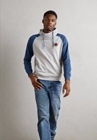 Sudadera gris y marino con mangas largas, bolsillo frontal y acento de logo. Combinada con pantalones vaqueros azules. Modelo de pie contra un fondo liso.