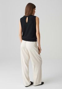 Top senza maniche nera con texture e dettaglio a chiave sul retro, abbinato a pantaloni bianchi larghi e scarpe nere.