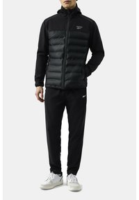 Czarna kurtka puchowa z wypełnieniem z pierza z przodu, zapięcie na zamek, wysoki kołnierz i elastyczne mankiety, zestawiona z czarnymi spodniami i białymi sneakersami.