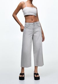 Femme portant un jean large gris clair raccourci, un débardeur assorti raccourci, et des sandales à plateforme noires sur un fond uni.