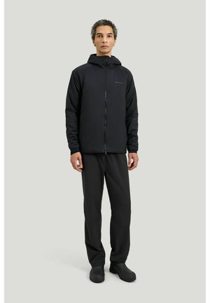 Mies seisoo päällään musta hupullinen vetoketjullinen takki, mustat housut ja mustat saappaat vaaleanharmaata taustaa vasten.