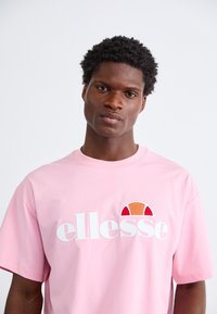 Rozā īsām piedurknēm T-krekls ar baltu "ellesse" zīmolu un krāsainu logo plāksteri oranžā un sarkanā krāsā. Gluda auduma tekstūra.