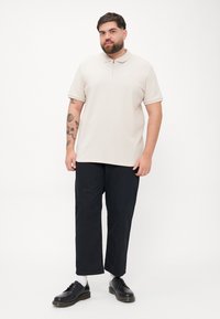 Beige polo T-shirt med krave og frontlynlås, parret med sorte bukser med vidde og sorte sko. Simpelt design og tekstureret stof.