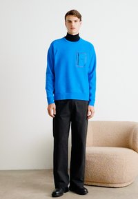 Felpa blu brillante con scollatura a girocollo e tasca sul petto, abbinata a pantaloni neri. Tessuto liscio e vestibilità rilassata.