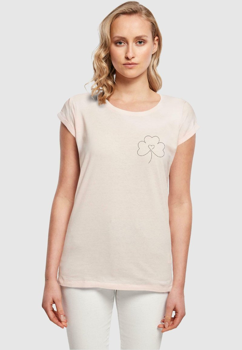 Lys rosa bomull t-skjorte med rullede ermer, rund hals, og et lite svart brodert kløverdesign på venstre bryst.