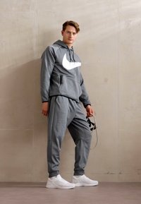 Felpa grigia in pile con un grande logo Nike bianco, pantaloni sportivi grigi coordinati e scarpe da ginnastica bianche. L'outfit presenta una vestibilità comoda.
