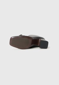 Pantof cu toc negru, cu un superior din piele netedă, vârf rotund și toc pătrat. Prezintă o talpă maronie cu o textură subtilă și detalii metalice.