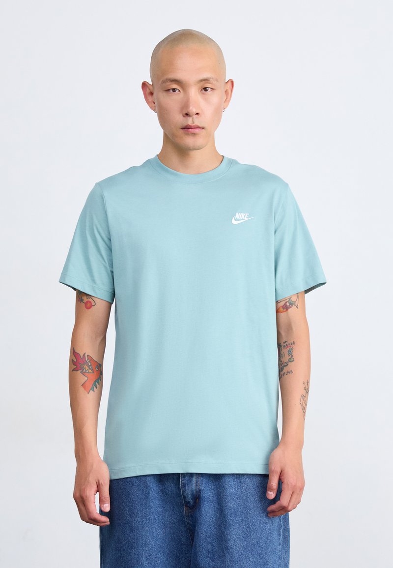 Άνδρας με ξυρισμένο κεφάλι που φοράει ανοιχτό μπλε μπλούζα Nike και μπλε τζιν, εκδηλώνοντας πολύχρωμα τατουάζ στους δύο μπροστινούς βραχίονες, που στέκεται μπροστά σε λευκό φόντο.