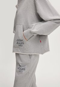 Szary dres składający się z krótkiej kurtki z przednią kieszenią. Materiał jest miękki; obie części mają nadruk "JOOP! JEANS CLUB" w ciemnoszarym kolorze.