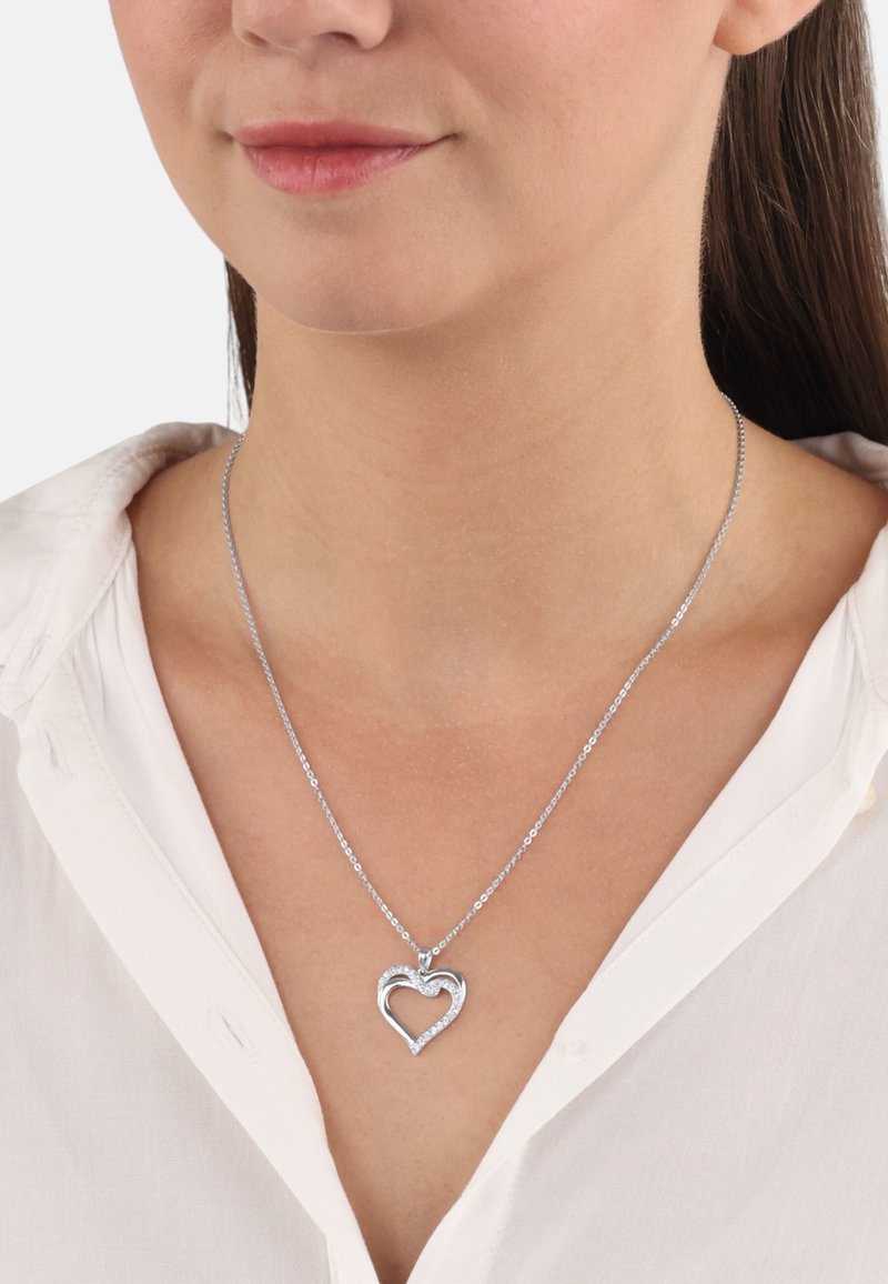 Collana d'argento con pendente a forma di cuore caratterizzato da un design testurizzato e accenti di diamante, poggiata su una camicetta bianca.