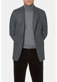 Blazer in lana grigia con rever a forma di intaglio, due tasche frontali e chiusura con un solo bottone, indossato sopra un dolcevita grigio chiaro, abbinato a pantaloni in velluto a coste marrone.