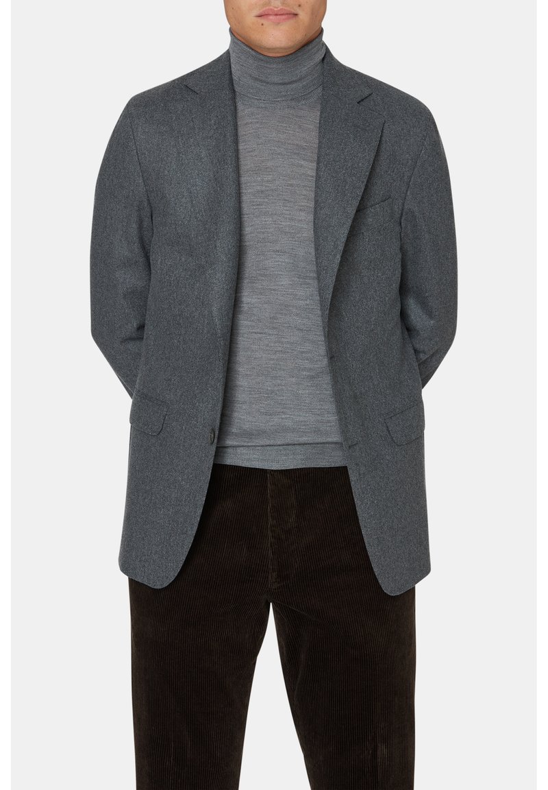 Blazer in lana grigia con rever a forma di intaglio, due tasche frontali e chiusura con un solo bottone, indossato sopra un dolcevita grigio chiaro, abbinato a pantaloni in velluto a coste marrone.