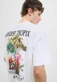 Hvid bomulds T-shirt med et farverigt tropisk grafikprint på ryggen med teksten "PARADISE TROPIC" og livlige planteillustrationer.