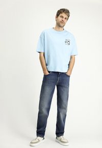 Camiseta oversized de color azul claro con un gráfico en el pecho, combinada con jeans rectos de color azul oscuro y zapatillas blancas con detalles grises.