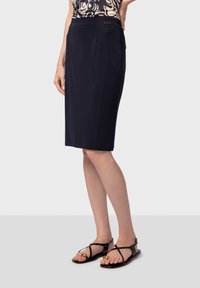 Jupe crayon bleu marine, longueur genou, avec une texture lisse, dotée de plis sur le devant et d'un accent métallique à la taille. Associée à des sandales noires.