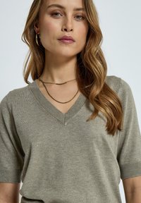 Maglione verde oliva chiaro a V con maniche corte, colletto e polsini a coste. Tessuto morbido, design minimalista, collana in oro a catena stratificata.