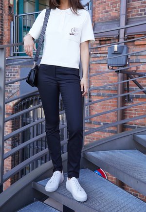 Mujer de pie en unas escaleras metálicas, vestida con una camiseta tipo polo blanca, pantalones negros, zapatillas blancas y llevando un bolso de hombro negro con una correa estampada.