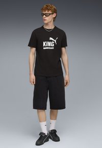 Camiseta de algodón negra con el texto en blanco "KING MANCHESTER CITY" y el logo de Puma, combinada con pantalones cortos de mezclilla negros y zapatillas negras.