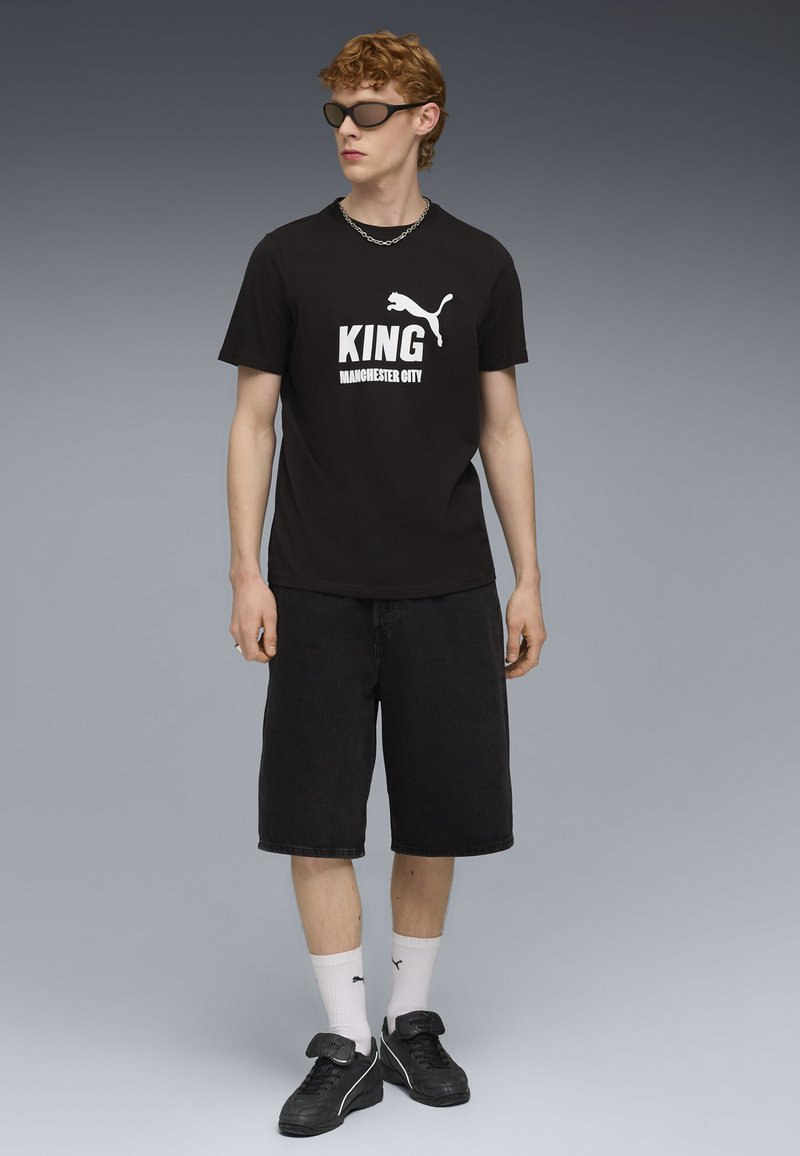 Camiseta de algodón negra con el texto en blanco "KING MANCHESTER CITY" y el logo de Puma, combinada con pantalones cortos de mezclilla negros y zapatillas negras.