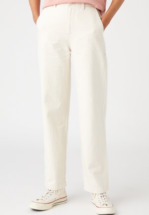 Persoon draagt ecru rechte broek met voorkanten zakken en witte lage sneakers met rode en blauwe strepen.