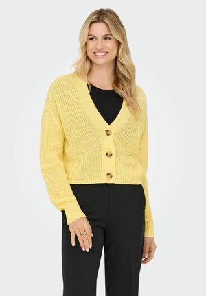 Yellow knitted cardigan with a v-neck, three large buttons, and long sleeves; cropped fit and ribbed cuffs; worn over a black top.