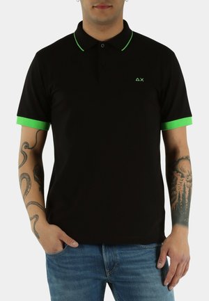 Uomo che indossa una polo nera con colletto e bordi delle maniche verdi, jeans blu e tatuaggi visibili sulle braccia, con le mani rilassate lungo i fianchi.