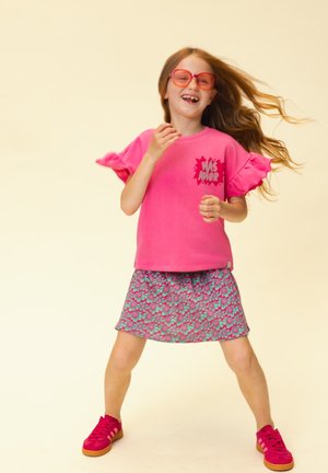 Jong meisje met lang haar, draagt een rode bril, een roze shirt met volantmouwen, een bloemenrok en rode sneakers, glimlacht en staat met gespreide benen.