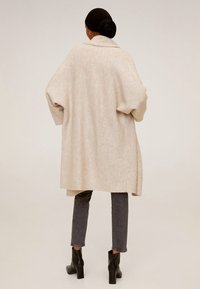 Femme portant un long cardigan beige oversize, un jean slim noir, des bottes noires à la cheville et un foulard noir, se tenant debout sur un fond uni.