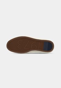 Sole of beige shoe with brown rubber bottom showing "1973" pattern and navy blue branded heel patch on a white background.