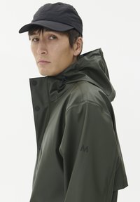 Olivgrön vattentät jacka med huva, prydd med svarta tryckknappar och en texturerad logga på ärmen. Bärs med en svart keps.