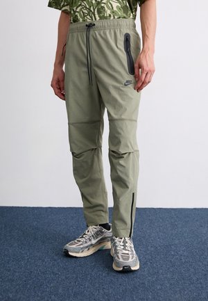 Mann trägt olivgrüne Nike-Jogginghose mit Reißverschlusstasche und graue Nike-Laufschuhe und steht auf einem blauen Teppich.