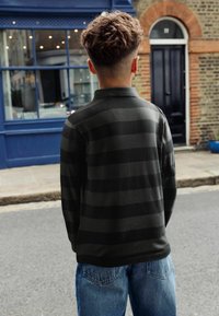 Sortstribet pullover med krave, lavet af et blødt materiale. Bæres med løstsiddende blå denimjeans. Baggrunden viser en blå butik.