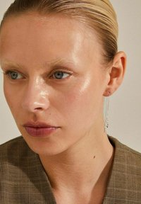 Strieborné visací náušnice s jedinečným špirálovým dizajnom, s elegantným reťazcom a malými akcentmi z drahokamov. Model má na sebe hnedý károvaný blejzer.