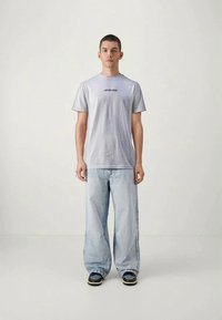 Jovem homem de frente, vestindo uma t-shirt cinza claro da Santa Cruz, jeans largos azul claro e ténis pretos, num fundo simples.