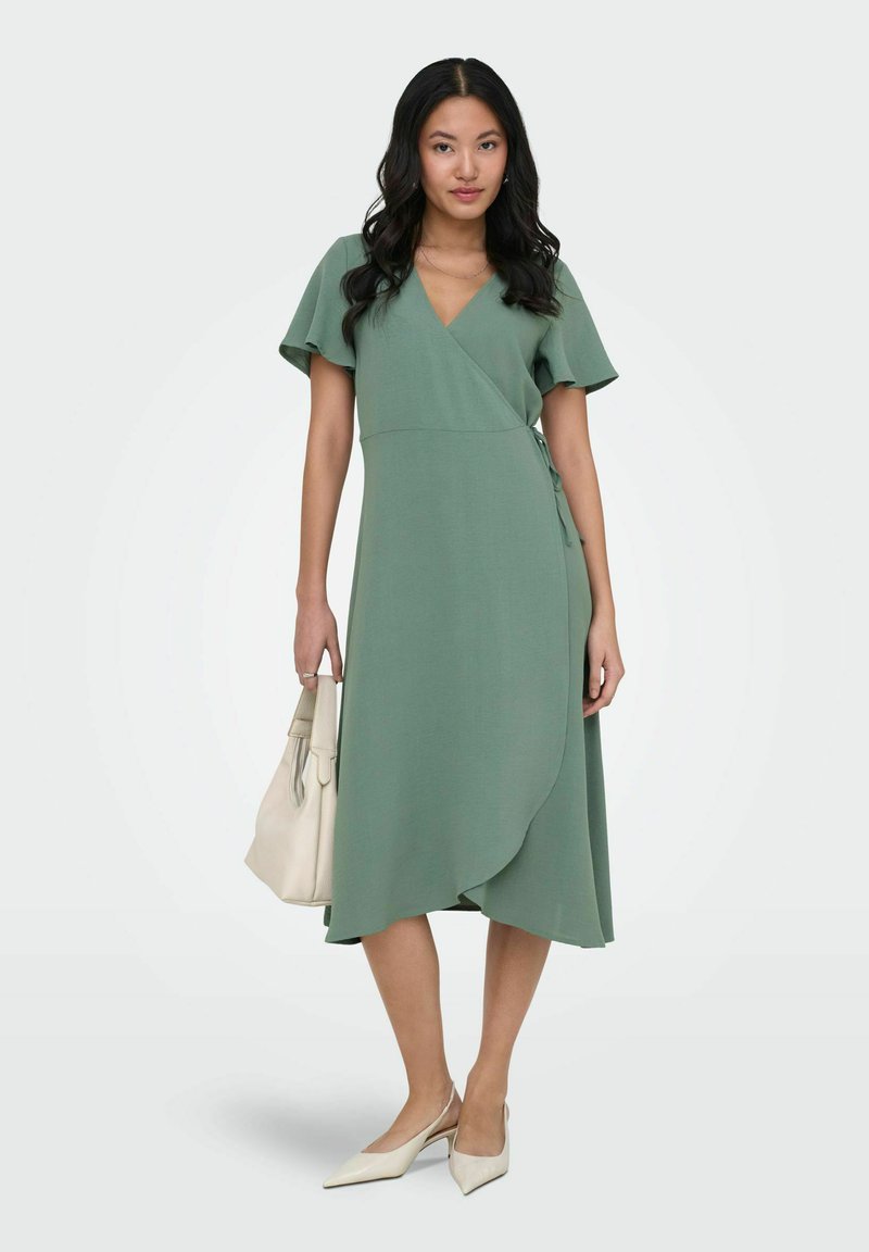 Femme portant une robe portefeuille vert sauge à manches volantées, tenant un sac à main crème et chaussée de talons slingback crème à bouts pointus.