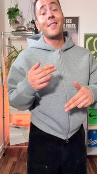 Jeune personne en sweat à capuche gris faisant un geste avec les mains dans une pièce avec des plantes, des étagères en métal et des affiches murales colorées.