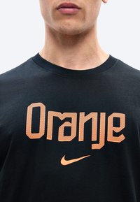 Κοντινό πλάνο ενός ατόμου που φοράει μαύρο πουκάμισο με το κείμενο "Oranje" και ένα πορτοκαλί λογότυπο Nike Swoosh από κάτω.