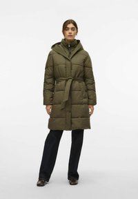 Ženska stoji v olivno zeleni puhovki z pasom in kapuco, temnih kavbojkah in rjavih čevljih, pred enobarvno belo ozadje.