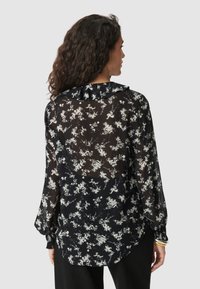 Black sheer blouse featuring a floral pattern in white, long sleeves, a ruffled collar, and a longer back hem. Soft, lightweight material.