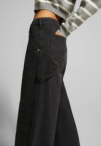 Person trägt schwarze Jeans mit hohem Bund und weitem Bein, mit Seitentasche, Hand in der Tasche, und einen kurz geschnittenen grauen und weißen gestreiften Pullover.