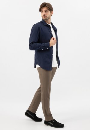 Uomo con camicia blu navy abbottonata sopra una t-shirt bianca, pantaloni marroni e scarpe nere, in piedi con un piede avanzato contro uno sfondo semplice.