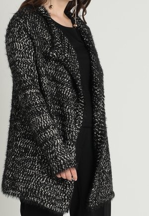 Femme portant un long cardigan en maille texturée noir et blanc sur un haut noir et un pantalon noir, sur un fond neutre.