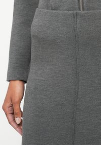 Robe grise à manches longues, en tissu tricot doux. Présente une fermeture éclair visible et une poche avant. Texture lisse avec des coutures subtiles.