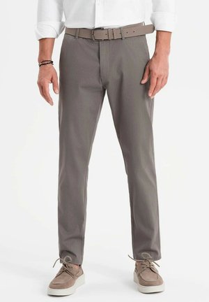 Pantalones grises con un acabado texturizado, corte recto y un cinturón a juego. Llevado con zapatos de ante marrón claro. Camisa blanca de botones.