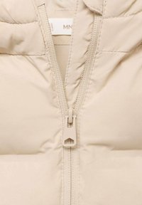Beige Pufferjacke mit einem Reißverschluss vorne. Sie hat eine gesteppten Textur, einen weichen Stoff und einen normalen Reißverschluss-Zieher in der Mitte.