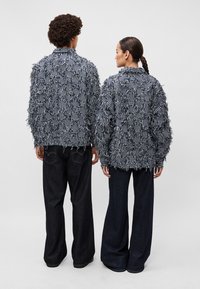 Adītas, pārmērīgas jakas jūras spēku un baltā svītru rakstā ar frills detaļām, uzvilktas virs tumšiem denima biksēm. Aizmugures skats uz diviem modeļiem.