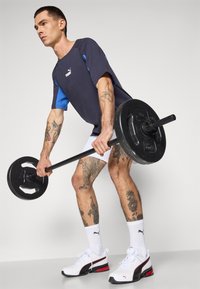 Zwarte halterstang vastgehouden door een persoon in een marineblauw en blauwe T-shirt, witte shorts en witte sneakers met rode accenten; zichtbare tatoeages op de benen.