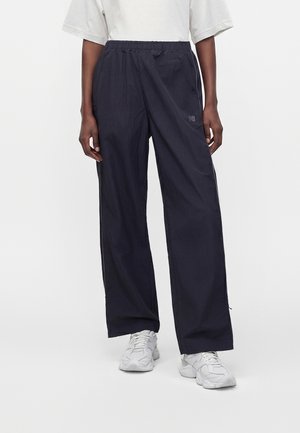 Person iført løstsiddende sorte træningsbukser og hvide sneakers, stående mod en ensfarvet hvid baggrund.